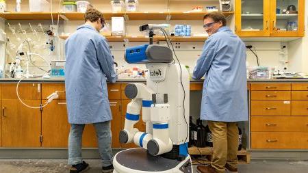 Two chemistry graduate students wearing in lab coats work at a lab bench while an automaton robot with a single, long arm prepares to work near them.